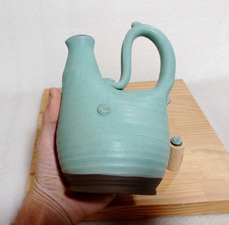 May include: A teal blue ceramic pitcher with a unique, curved handle. The pitcher has a textured surface and a dark brown base.