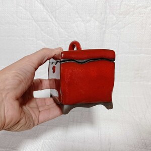 May include: A small, red ceramic box with a handle on top. The box has a dark brown glaze and a slightly textured surface. The box is sitting on a white surface.