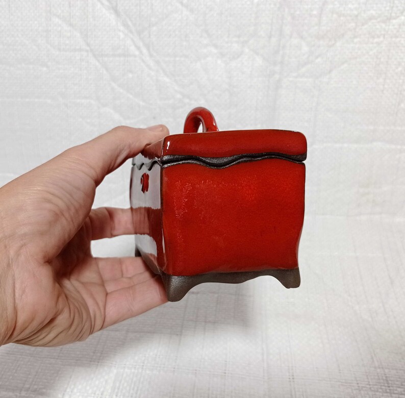 May include: A small, red ceramic box with a handle on top. The box has a dark brown glaze and a slightly textured surface. The box is sitting on a white surface.