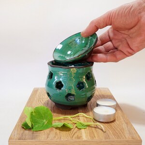 May include: A teal ceramic oil burner with a removable lid and decorative cutouts. The burner is sitting on a wooden surface with green leaves and two white candles.