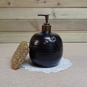 May include: A black ceramic soap dispenser with a brass pump and a swirl design. The dispenser is sitting on a white doily.