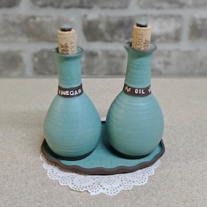 May include: Two teal ceramic oil and vinegar bottles with cork stoppers and brown labels that say "Oil" and "Vinegar". The bottles are on a teal tray with a brown rim.