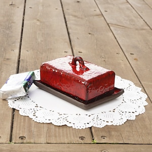 May include: Red ceramic butter dish with a lid and a handle. The dish is sitting on a white doily on a wooden surface.