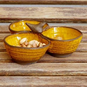 May include: A three-sectioned ceramic serving dish with a bird-shaped handle. The dish is glazed in a yellow-brown color and has a textured finish. The dish is filled with pistachios.