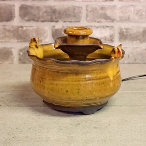 May include: A small, ceramic fountain with a tiered design. The fountain is glazed in a yellow and brown color scheme. The top tier has a small, round opening for water to flow through. The fountain is sitting on a gray surface.