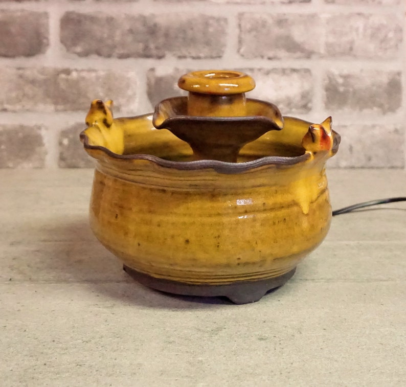 May include: A small, ceramic fountain with a tiered design. The fountain is glazed in a yellow and brown color scheme. The top tier has a small, round opening for water to flow through. The fountain is sitting on a gray surface.