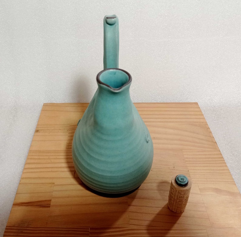 May include: A teal blue ceramic pitcher with a curved spout and a wide base. The pitcher is sitting on a wooden surface.