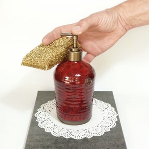May include: A red ceramic soap dispenser with a gold pump top. The dispenser is sitting on a white doily on a gray surface. A gold sponge is in the background.
