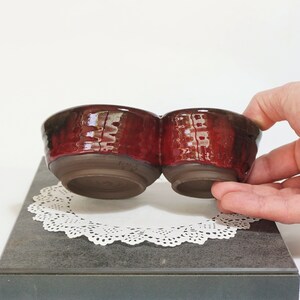 May include: A red ceramic double dish with a glossy finish. The dish is made of two connected bowls, each with a rounded shape. The dish is sitting on a white doily.