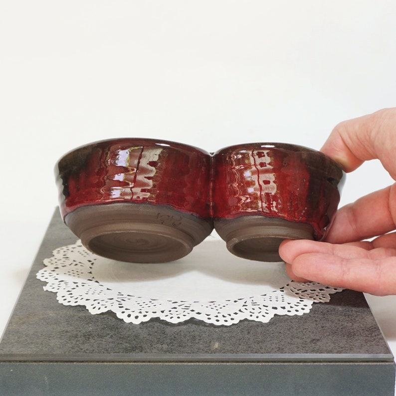May include: A red ceramic double dish with a glossy finish. The dish is made of two connected bowls, each with a rounded shape. The dish is sitting on a white doily.