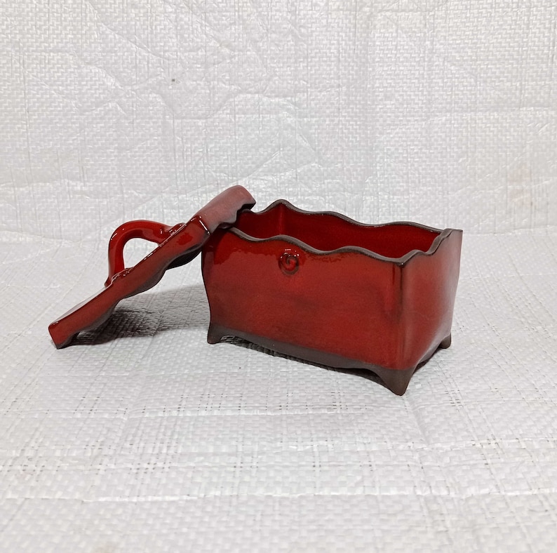 May include: A red ceramic box with a lid. The box has a wavy edge and a handle on the lid. The box is sitting on a white surface.