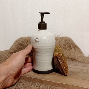 May include: White ceramic soap dispenser with a black rim and a brown pump. The dispenser is sitting on a wooden surface with a brown burlap background.