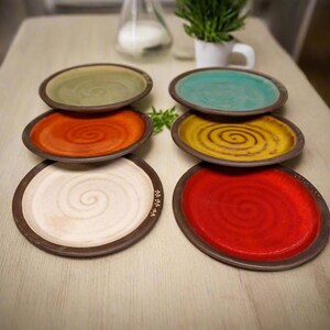 May include: Six ceramic plates with a spiral design in various colors, including red, orange, yellow, green, and white. The plates are arranged in a row on a light wood surface.