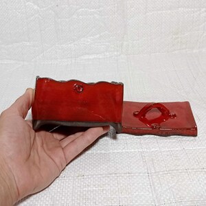 May include: A red ceramic box with a lid. The box has a wavy edge and a small handle on the lid. The box is sitting on a white surface.