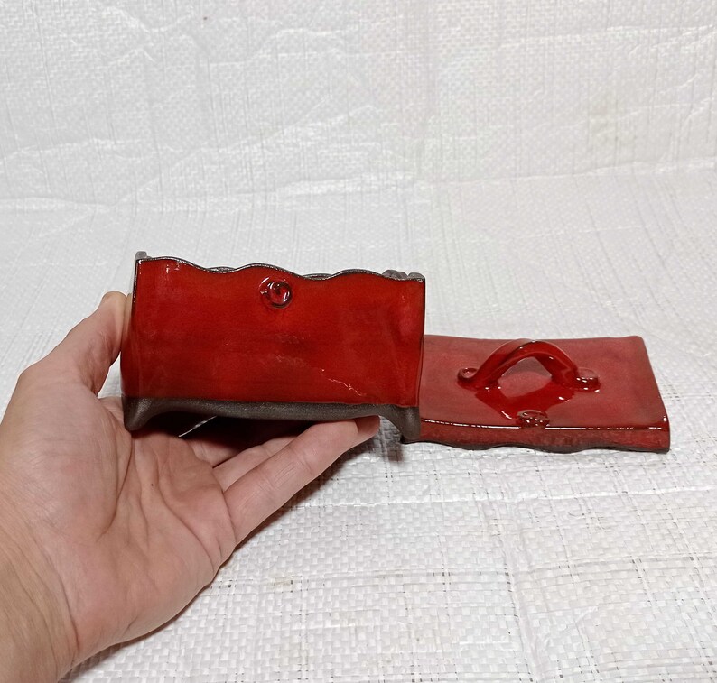 May include: A red ceramic box with a lid. The box has a wavy edge and a small handle on the lid. The box is sitting on a white surface.