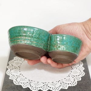 May include: A set of two teal green ceramic bowls with a shared base. The bowls are glazed with a speckled finish and have a rustic brown rim.