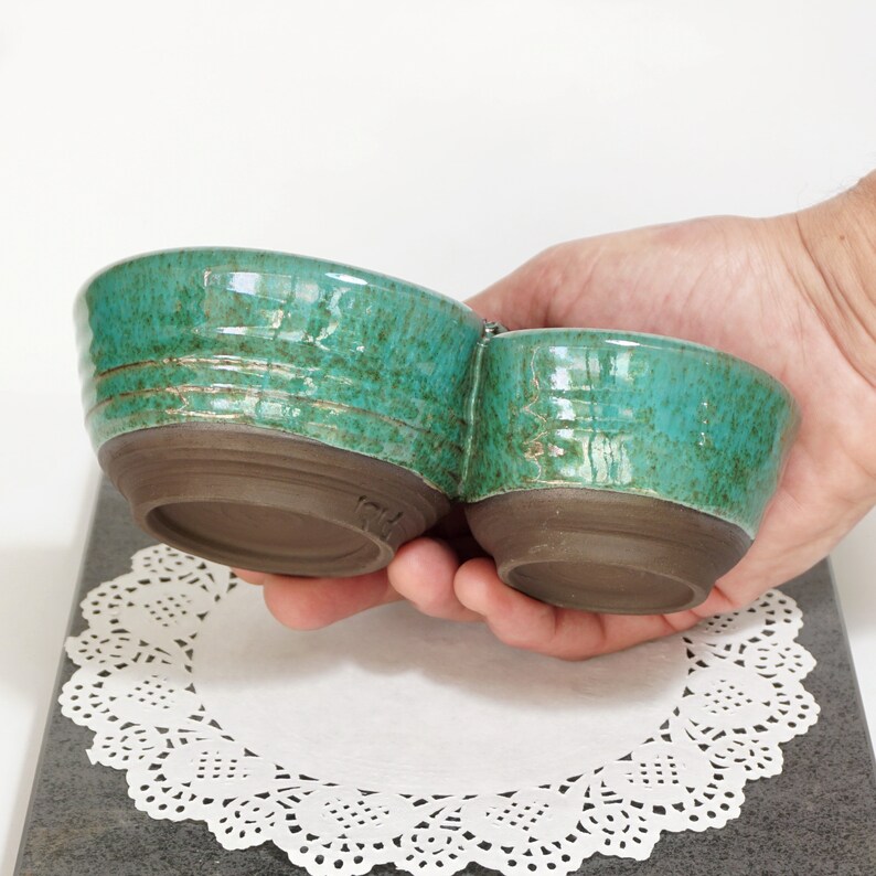 May include: A set of two teal green ceramic bowls with a shared base. The bowls are glazed with a speckled finish and have a rustic brown rim.