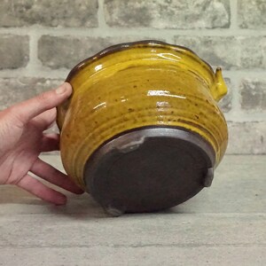 May include: A yellow ceramic planter with a brown base and a slightly flared rim. The planter has a unique, slightly asymmetrical shape.