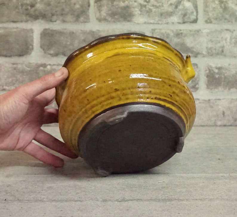 May include: A yellow ceramic planter with a brown base and a slightly flared rim. The planter has a unique, slightly asymmetrical shape.