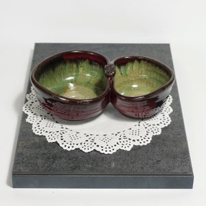 May include: A double-sided ceramic serving dish with a red and green glaze. The dish has two bowls connected by a small handle.