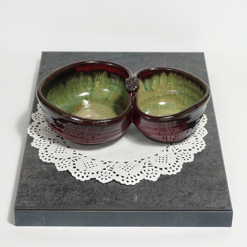 May include: A double-sided ceramic serving dish with a red and green glaze. The dish has two bowls connected by a small handle.