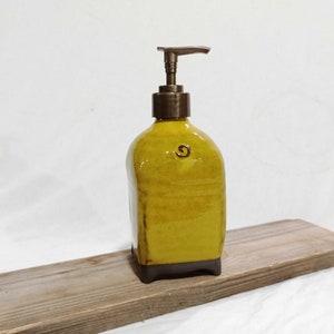 May include: A yellow ceramic soap dispenser with a brown base and a brass pump. The dispenser is sitting on a wooden surface.