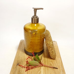 May include: A yellow ceramic soap dispenser with a bronze pump and a swirl design. The dispenser is sitting on a wooden surface with a gold sponge.