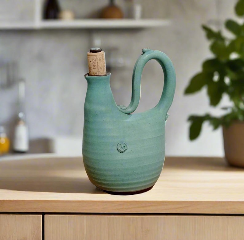 May include: A teal blue ceramic pitcher with a unique curved handle and a cork stopper. The pitcher has a textured surface and a subtle swirl design.