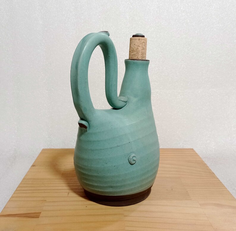 May include: A teal blue ceramic bottle with a cork stopper and a unique, curved handle. The bottle has a textured surface and a brown rim.