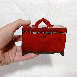 May include: A small, red ceramic box with a handle. The box has a black glaze and a decorative swirl on the front.