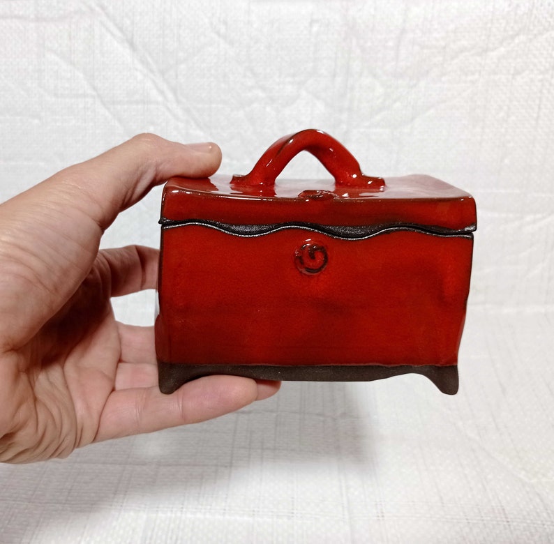 May include: A small, red ceramic box with a handle. The box has a black glaze and a decorative swirl on the front.