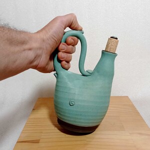 May include: A teal blue ceramic pitcher with a cork stopper and a handle. The pitcher has a textured surface and a brown base.
