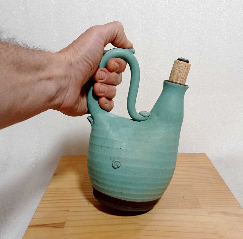 May include: A teal blue ceramic pitcher with a cork stopper and a handle. The pitcher has a textured surface and a brown base.