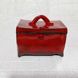 May include: A red ceramic box with a handle on the top. The box has a glossy finish and a decorative design on the front.