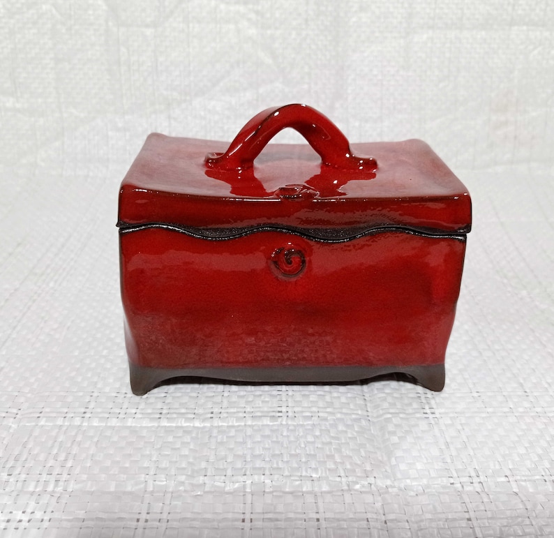 May include: A red ceramic box with a handle on the top. The box has a glossy finish and a decorative design on the front.