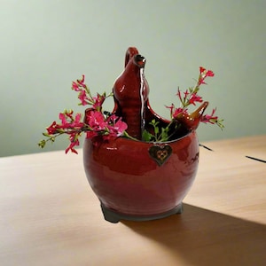 Könnte beinhalten: Eine rote Keramikfontäne mit einem Vogelschnabel und einem Herzdesign. Die Fontäne ist mit Wasser gefüllt und hat eine kleine Pflanze, die darin wächst. Rosafarbene Blumen sind um die Fontäne herum angeordnet.