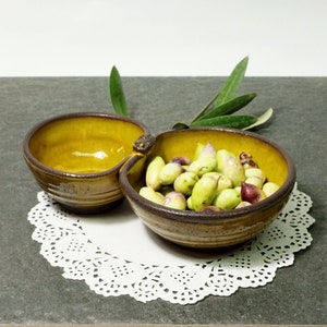 May include: A brown and yellow ceramic serving dish with two sections. The larger section is filled with green olives. The dish is sitting on a white doily.