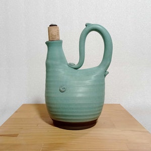 May include: A light blue ceramic pitcher with a cork stopper and a unique handle. The pitcher has a textured surface and a brown base.