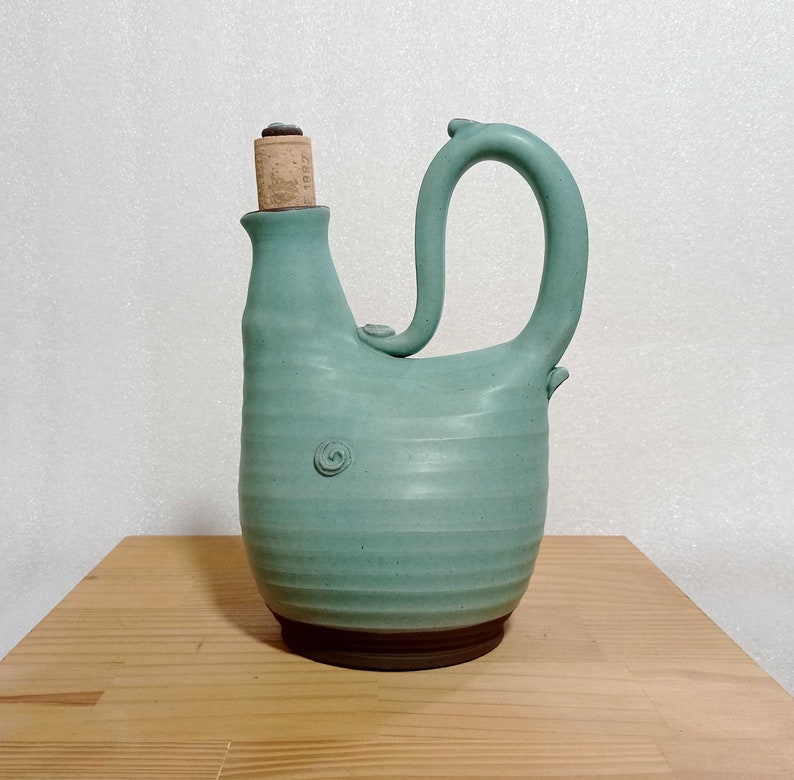 May include: A light blue ceramic pitcher with a cork stopper and a unique handle. The pitcher has a textured surface and a brown base.