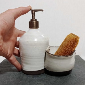 May include: White ceramic soap dispenser with a brown base and a metal pump. The dispenser is next to a white ceramic dish with a brown base, holding a brown sponge.
