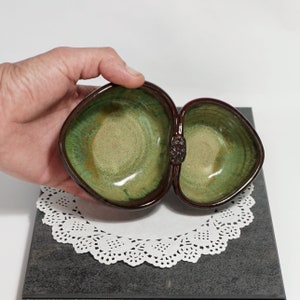 May include: A two-part ceramic dish with a green and brown glaze. The dish is heart-shaped and has a raised design on the side where the two parts connect.