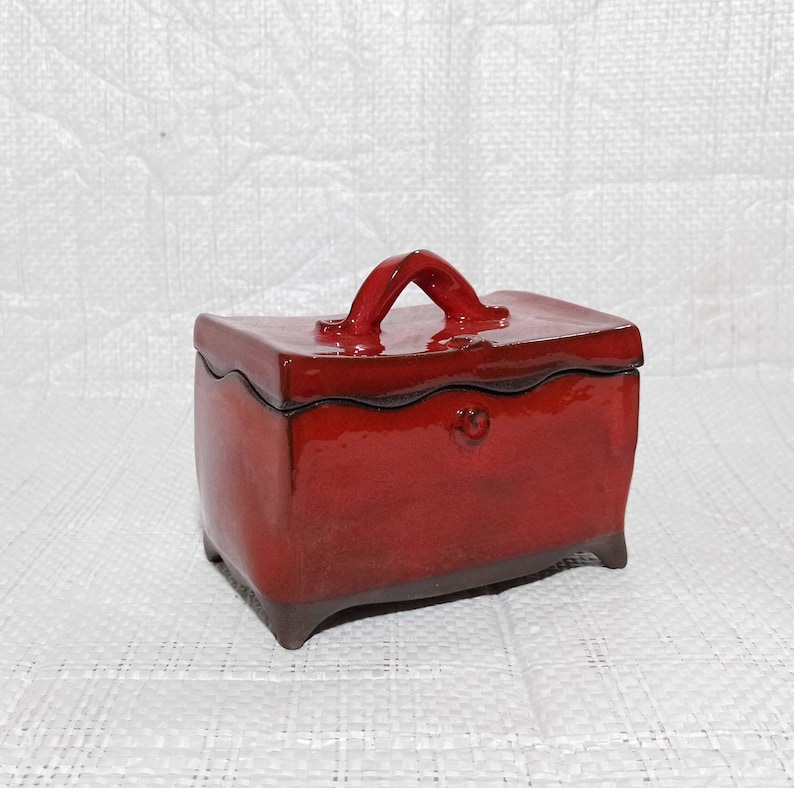 May include: A red ceramic box with a handle on the top. The box has a slightly rounded shape and sits on four small feet.