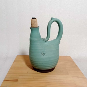 May include: A teal blue ceramic pitcher with a unique spout and handle. The pitcher has a cork stopper with the text "1882" on it.