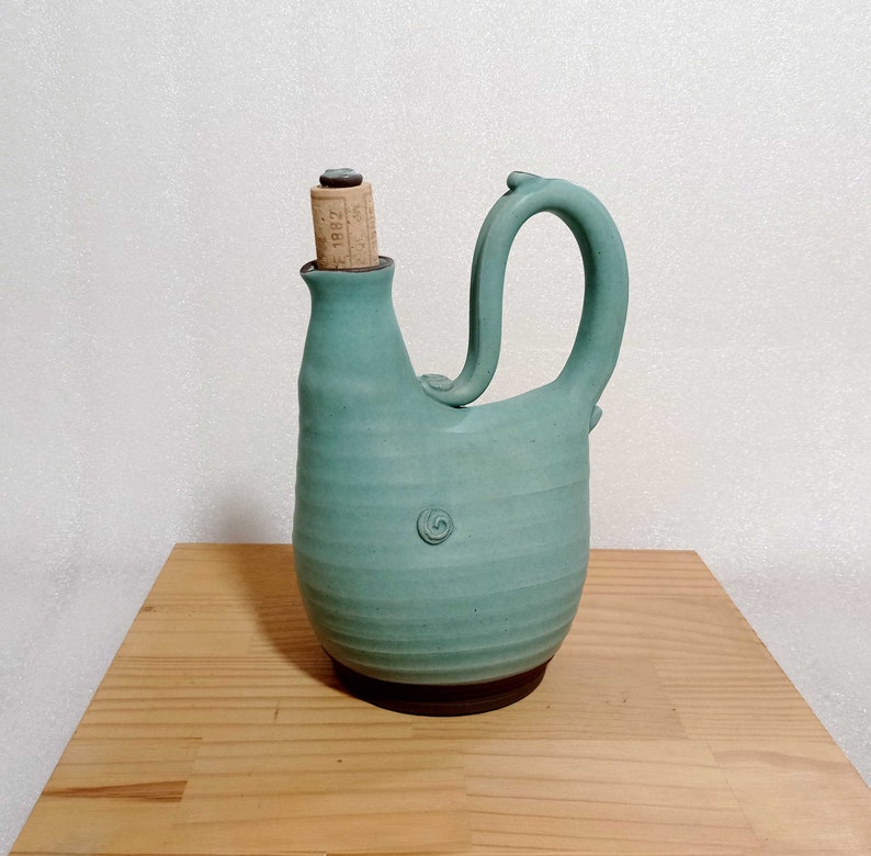 May include: A teal blue ceramic pitcher with a unique spout and handle. The pitcher has a cork stopper with the text "1882" on it.