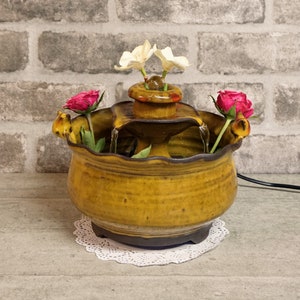 May include: A ceramic tabletop fountain with a three-tiered waterfall design. The fountain is decorated with a yellow glaze and features a brown base. The fountain is surrounded by white and pink flowers.