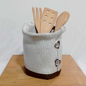 May include: White ceramic utensil holder with a brown rim and a textured surface resembling burlap. The holder has two brown swirl designs on the side and is filled with wooden kitchen utensils.