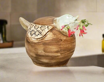 May include: A ceramic bird figurine with a light brown and white glaze. The bird has a small, open beak with a sprig of red flowers sticking out. The bird's body is decorated with a white floral pattern.