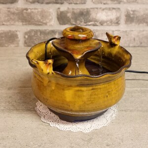May include: A ceramic tabletop fountain with a tiered design. The fountain is decorated with a brown glaze and has a rustic look. The fountain has a small, round basin at the top and a larger, round basin at the bottom. Water flows from the top basin to the bottom basin.