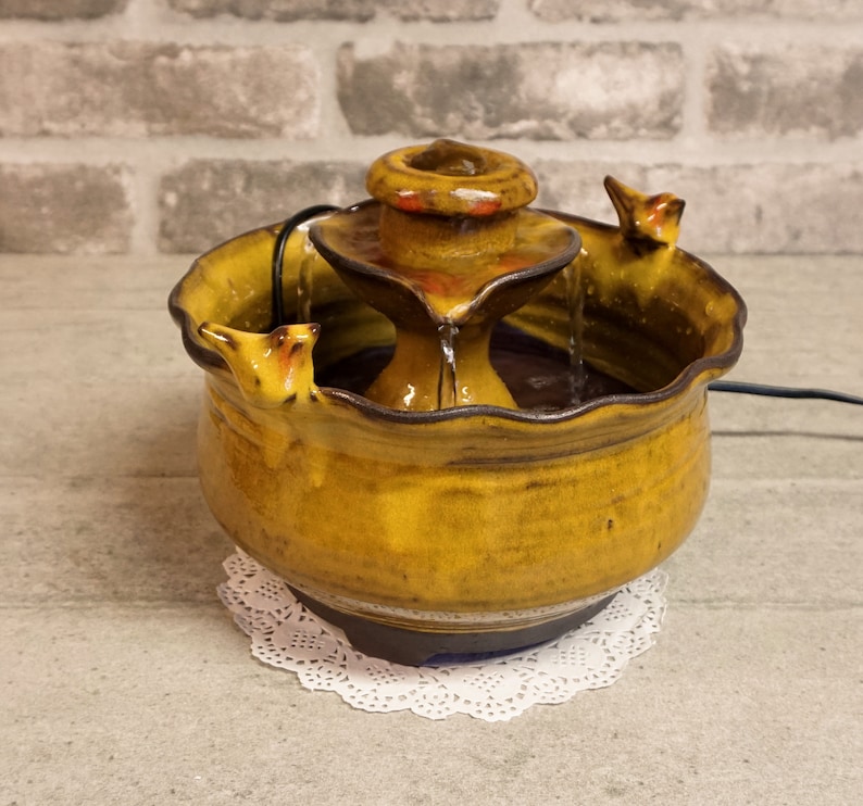 May include: A ceramic tabletop fountain with a tiered design. The fountain is decorated with a brown glaze and has a rustic look. The fountain has a small, round basin at the top and a larger, round basin at the bottom. Water flows from the top basin to the bottom basin.