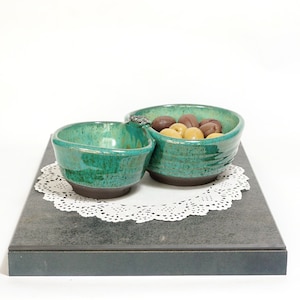 May include: A teal ceramic serving dish with two connected bowls. The dish is filled with olives and sits on a white doily on a black surface.